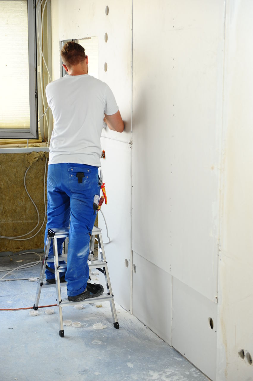 Person on a ladder applying paint to a wall in a room under renovation.